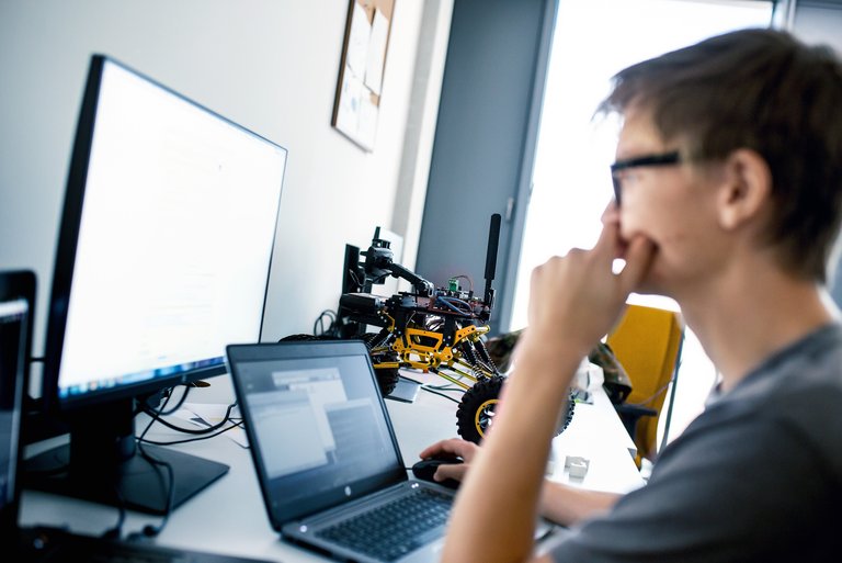 Ein junger Mann mit Brille sitzt vor einem Laptop und einem Bildschirm, während im Hintergrund ein Spielzeugrennauto steht