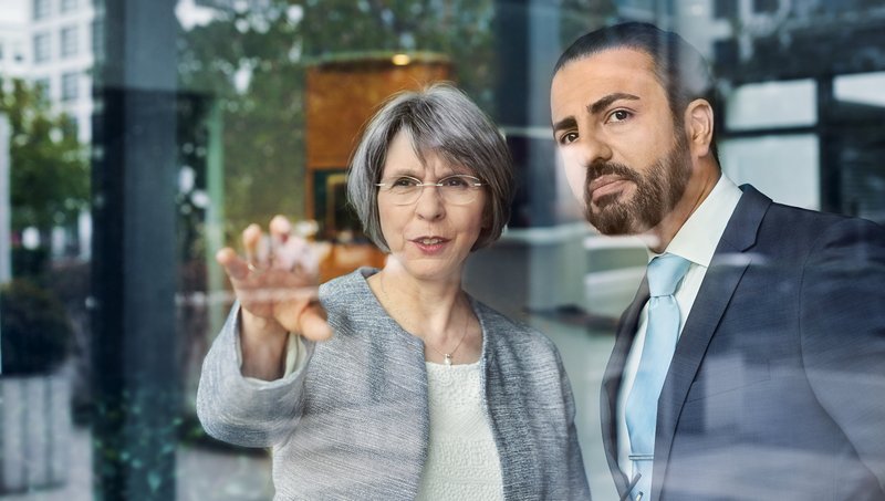 A woman with grey hair and glasses shows a man with a beard and a suit something in the distance behind a pane of glass