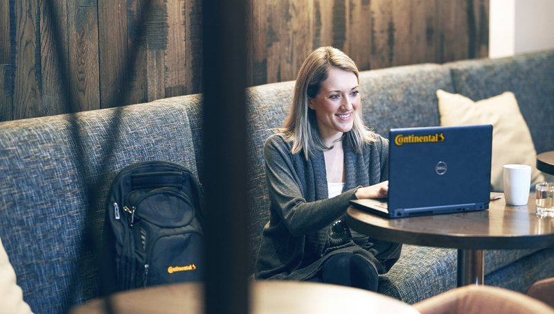 Eine Frau sitzt auf einem Sofa, neben ihr ein Rucksack mit Continental-Logo, und arbeitet lächelnd an einem Laptop mit Continental-Logo