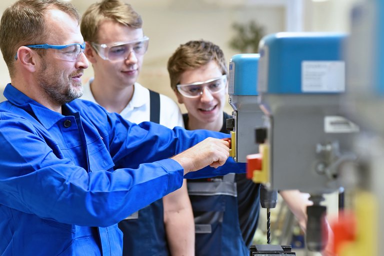 Ein Mann in Blaumann und Schutzbrille zeigt zwei Jungs in Latzhosen die Bedienung einer technischen Maschine