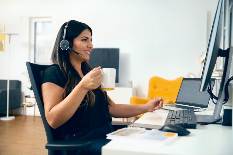 Eine junge Frau mit dunklen Haaren in schwarzem T-Shirt trägt ein Head-Set und hält einen Kaffee-Pot während Sie an ihrem Home Office Arbeitsplatz sitzt