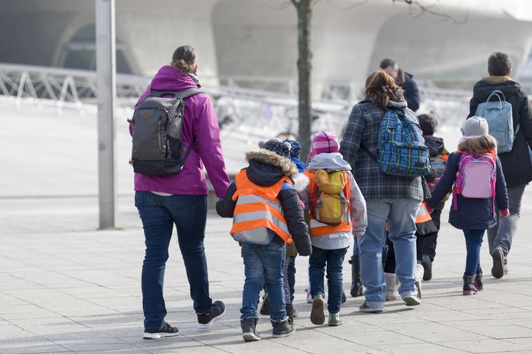 Eine Gruppe von Grundschulkindern und Betreuer:innen geht Hand in Hand auf ein futuristisches Gebäude zu