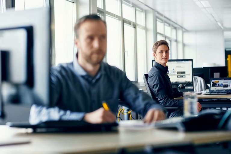 Ein an einem Schreibtisch im Hintergrund schaut auf einen Mann am Schreibtisch im Vordergrund