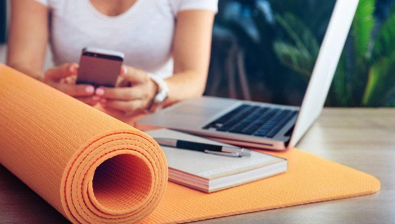 Eine halb zusammengerollte Yoga-Matte liegt mit einem Notizbuch, Stift und Laptop auf einem Tisch, während eine Frau im Hintergrund auf einem Handy tippt