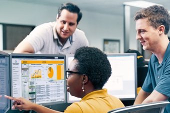 A middle-aged woman sits in front of three PC screens displaying statistics and shows two men something on the left display.