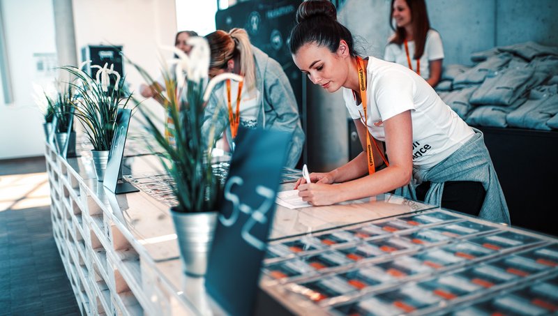 A young woman in a white T-shirt and with a lanyard around her neck is standing at a trade fair booth and writing something down on a piece of paper