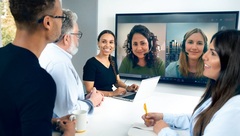 Zwei junge Frauen und zwei Männer stehen an einem Stehtisch und blicken auf einen Fernseher, auf dem zwei Frauen mit Headset in einer Videokonferenz abgebildet sind