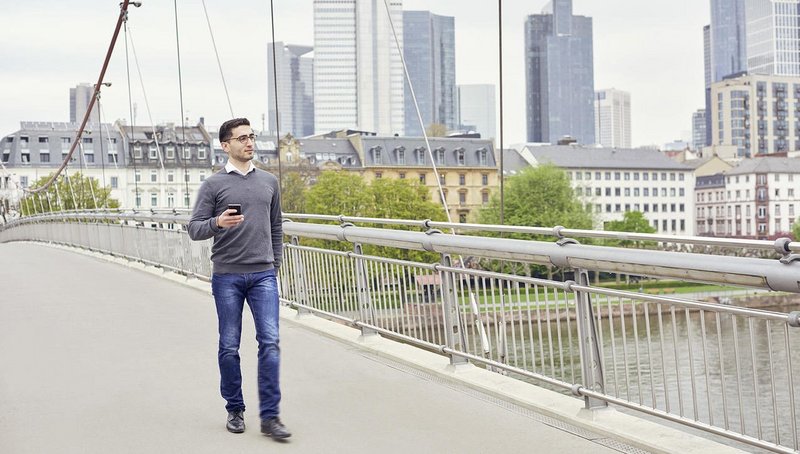 A young man is walking over a bridge in front of the skyline of Frankfurt, Germany