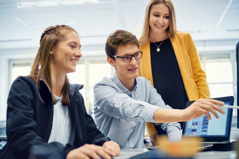 Ein junger Mann mit Brille zeigt zwei jungen Frauen etwas auf seinem PC