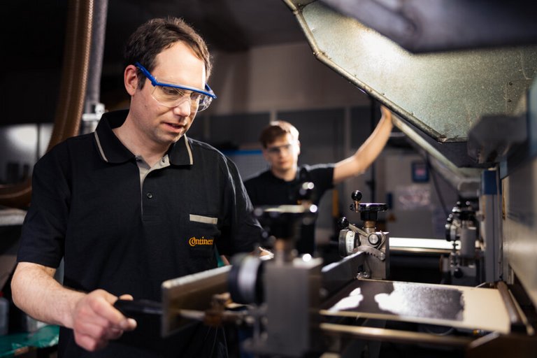 Ein junger Mann in schwarzem Polohemd und Schutzbrille steht in einem Labor und hebt mit einem Löffel eine braune Substanz aus einem Glas