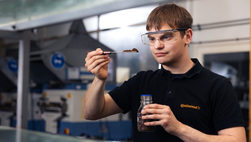 A young man in a black polo shirt and safety goggles standing in a laboratory and using a spoon to lift a brown substance out of a glass