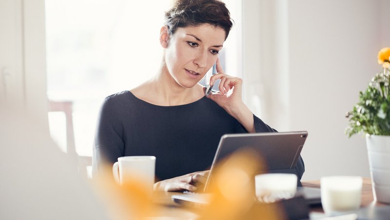 Eine Frau sitzt am Tisch und arbeitet von zu Hause mit ihrem Tablet und hält ihr Handy am Ohr