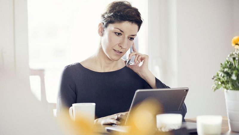 A woman sits at the table, working from home with the tablet in front of her and  the mobile phone to her ear