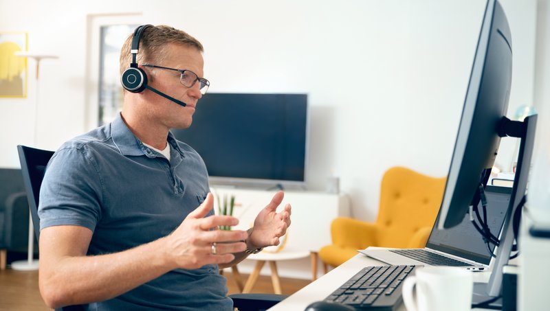 A man with glasses, polo shirt and headset sits gesticulating in front of a workplace