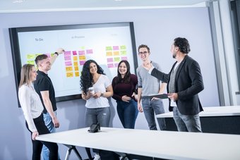 Drei junge Frauen und zwei junge Männer stehen vor einem White Board und blicken zu einem mittelalten Mann im Sakko, der die Inhalte des White Boards erklärt