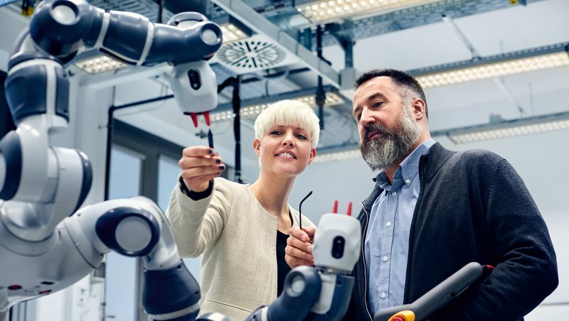 Két nő és egy férfi egy dolgozó robot előtt állnak egy gyárban