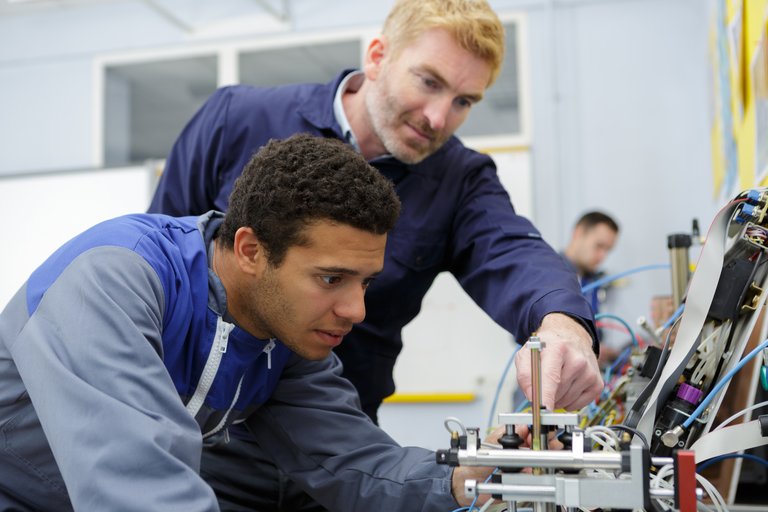 Ein junger Mann in Arbeitskleidung werkelt an einem technischen Gegenstand mit Kabeln, während ihm ein älterer Mann über die Schulter schaut und Tipps gibt