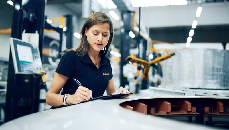 Eine junge Frau im Poloshirt steht in einer Fabrik und notiert etwas