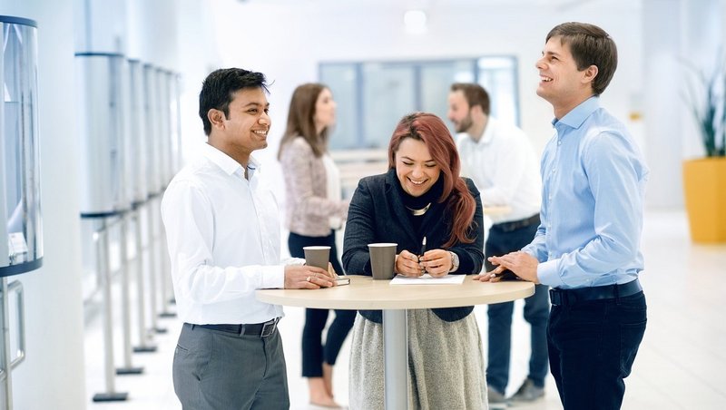 Zwei Männer und zwei Frauen in Business-Outfits gehen einen lichtdurchfluteten Flur mit bodentiefen Fensterscheiben entlang und lächeln sich an