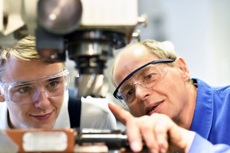 Ein älterer Mann in Blaumann und Schutzbrille zeigt einem Jungen mit Schutzbrille eine technische Maschine
