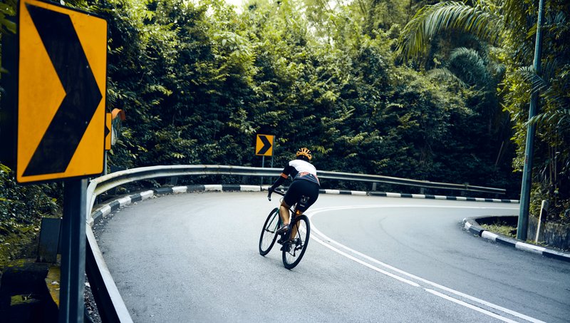 A racing cyclist turns into the bend of a dual carriageway bordered by a crash barrier, road signs trees and bushes