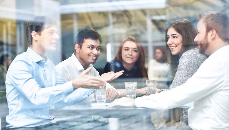 Three men and two women sit across from each other at a table and chat joyfully behind a pane of glass