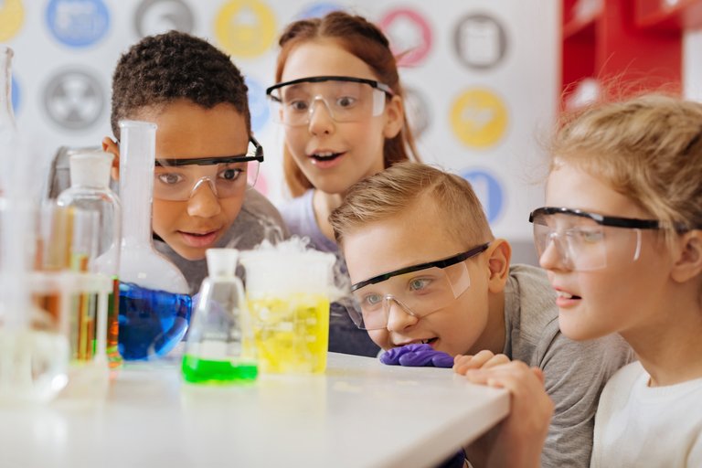 Zwei Mädchen und zwei Jungen im Grundschulalter tragen Sicherheitsbrillen und staunen über brodelnde bunte Reagenzgläser auf dem Tisch vor ihnen