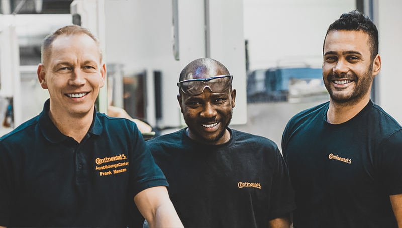 Drei Männer in schwarzen Continental-Poloshirts stehen in einer Fabrikhalle und lächeln