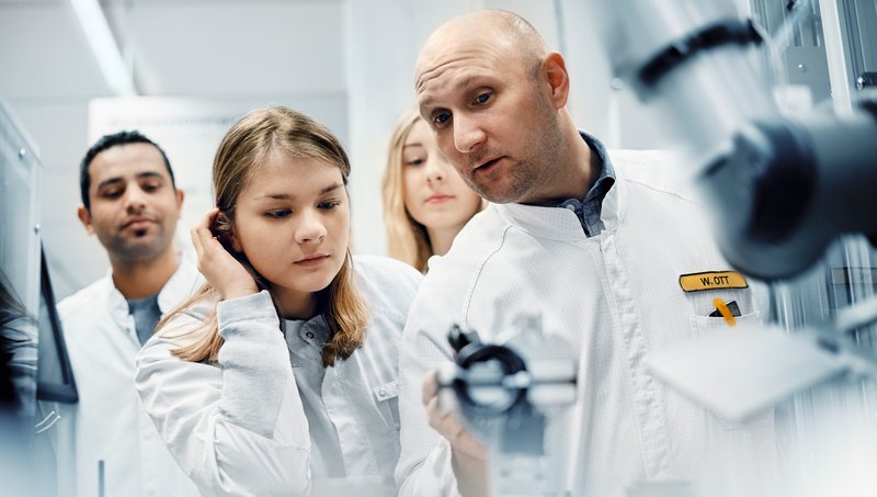 A man in a smock is explaining the functioning of a machine to a group of three young people