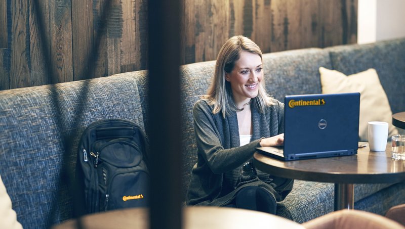 A woman sits on a sofa, next to her a backpack with Continental logo, and works smiling on a laptop with Continental logo