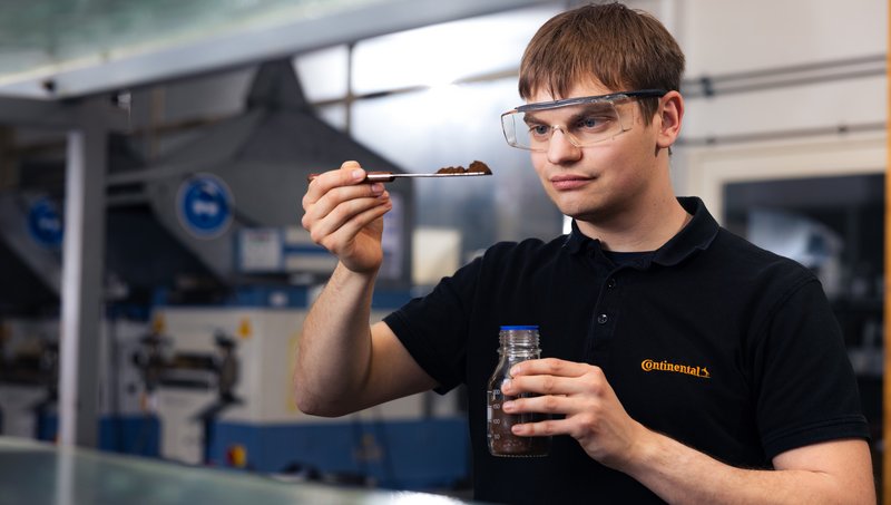 Ein junger Mann in schwarzem Polohemd und Schutzbrille steht in einem Labor und hebt mit einem Löffel eine braune Substanz aus einem Glas