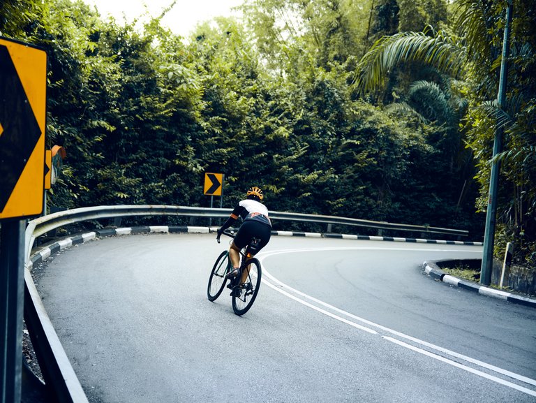 Ein Rennradfahrer biegt in die Kurve einer Schnellstraße ein, die von einer Leitplanke, Straßenschildern Bäumen und Büschen eingegrenzt wird <p>: Eine berufliche Auszeit für die Verwirklichung der persönlichen Träume nutzen und unkompliziert wiedereinsteigen.